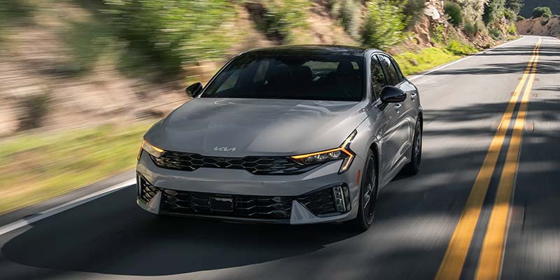 A sleek gray 2026 Kia K5 sedan driving on a curvy road surrounded by green trees.