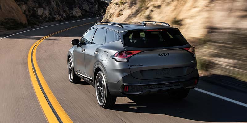 A sleek, silver Kia Sportage Hybrid drives along a winding mountain road, highlighted by bright yellow lines. The scene conveys a sense of adventure and freedom.