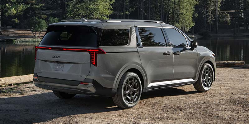 2026 Kia Carnival Hybrid parked beside a lake, showcasing its sleek gray design and modern rims.